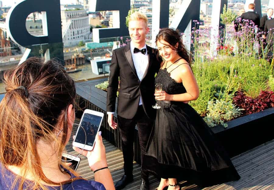 Two people, dressed in gala style, are standing on a roof terrace. A third person is taking a picture of them on their phone. The weather is sunny.