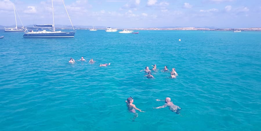 A group of people enjoy a swim in the ocean. There are boats in the distance.