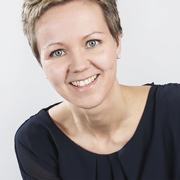 A woman with short brown hair smiles into the camera. It's a studio setting.