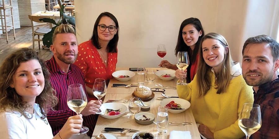 Six people sit a t a table in a restaurant. They are holding a glass of wine and are smiling at the camera.