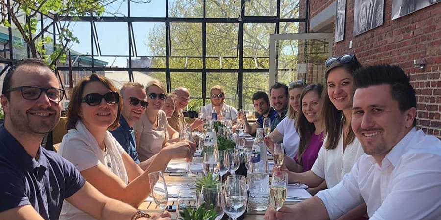 A group of approximately fifteen people sit at a long table and are having a drink. They are all smiling at the camera. They are wearing casual clothes.