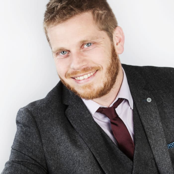 A man with dark blond hair, wearing a suit, smiles into the camera. It's a studio setting.