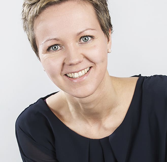 A woman with short brown hair smiles into the camera. It's a studio setting.