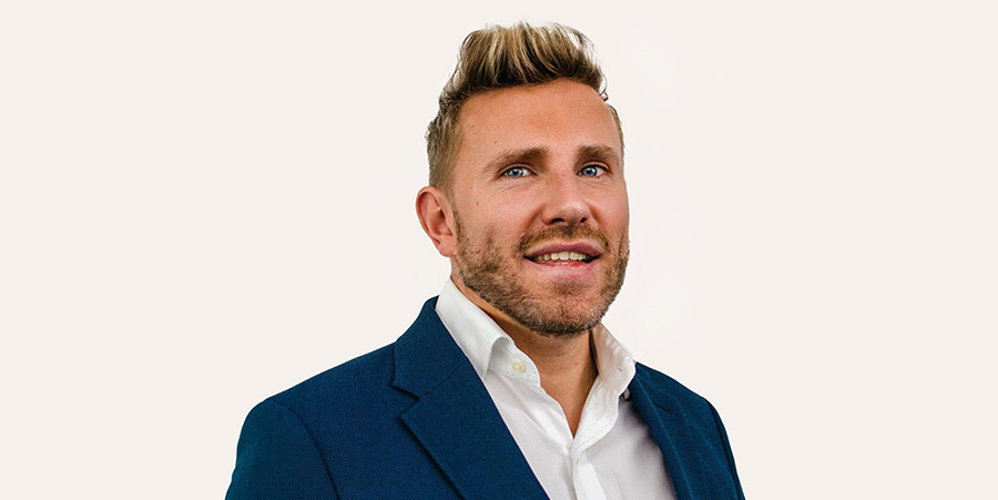 A man with blond hair, wearing a suit, smiles into the camera. The background is a white backdrop with the letters Hays working for your tomorrow.