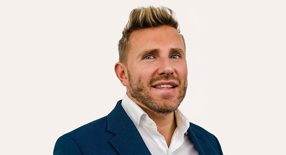 A man with blond hair, wearing a suit, smiles into the camera. The background is a white backdrop with the letters Hays working for your tomorrow.